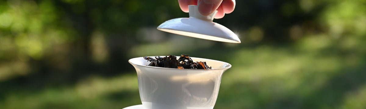 A hand lifting the lid of a white ceramic gaiwan tea bowl, showing loose-leaf tea inside, set on a rustic wooden table with a blurred green outdoor background.